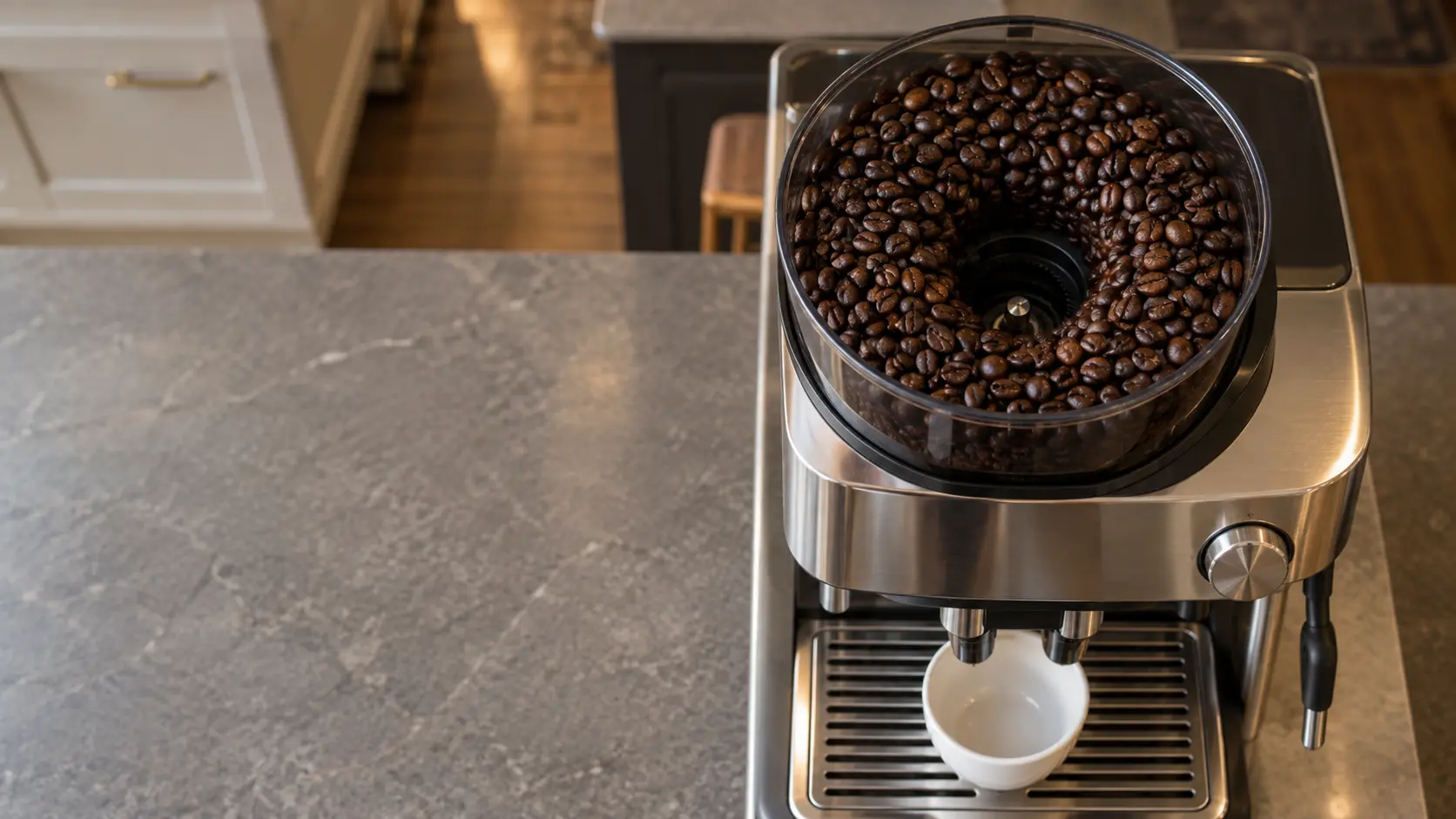 Top-down view into the open bean hopper of a stainless countertop coffee machine full of glossy dark coffee beans clumped around a hollow above the burrs, a white cup under the spouts at the lower edge of the frame, the coffee machine on a gray stone counter.